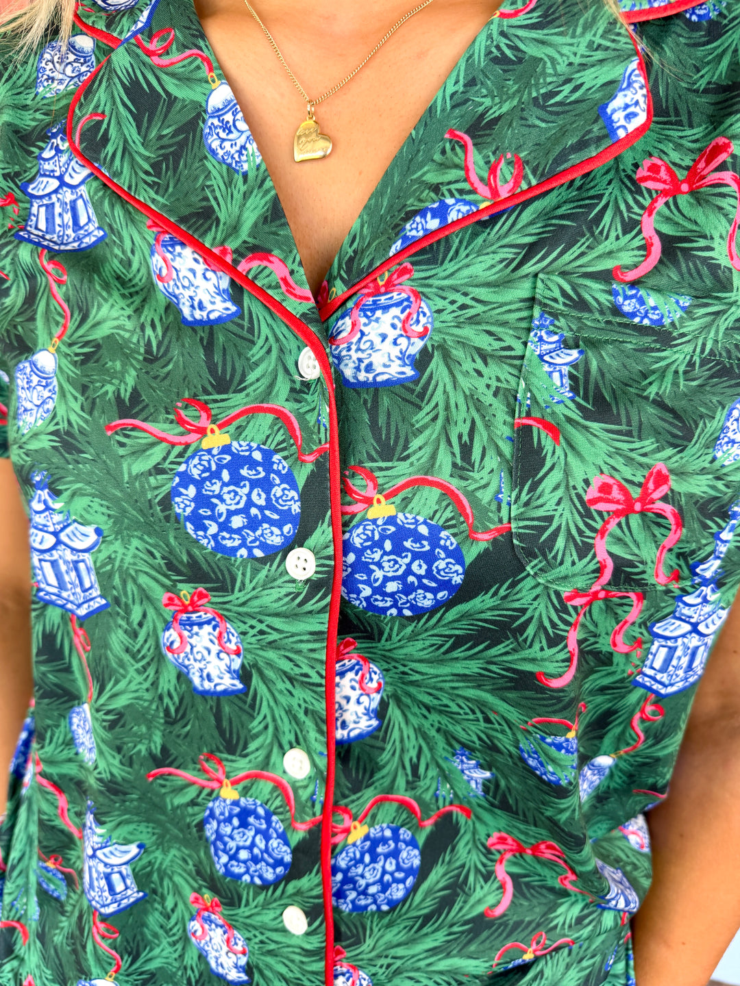 A close-up of a person wearing a green pajama top with a Christmas tree branch print, decorated with blue patterned ornaments, red bows, and red piping along the collar and front placket.