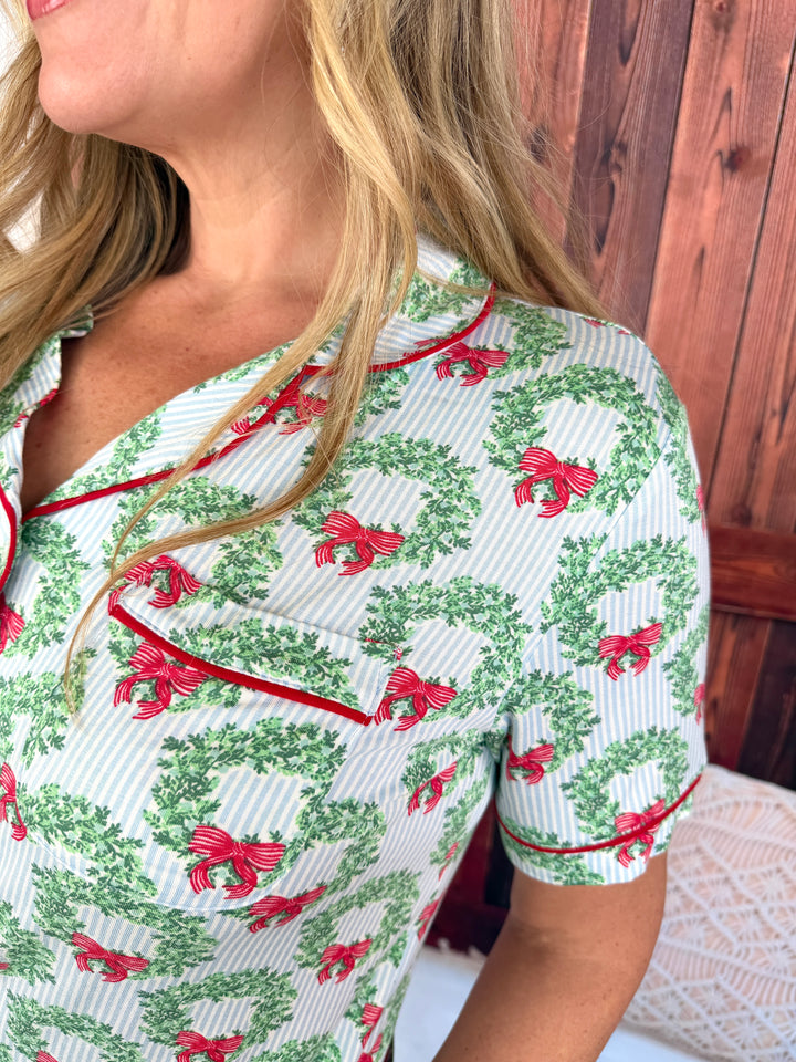 A close-up of a woman wearing a short-sleeve pajama top with a green wreath and red bow pattern on a white striped background, featuring red piping details.
