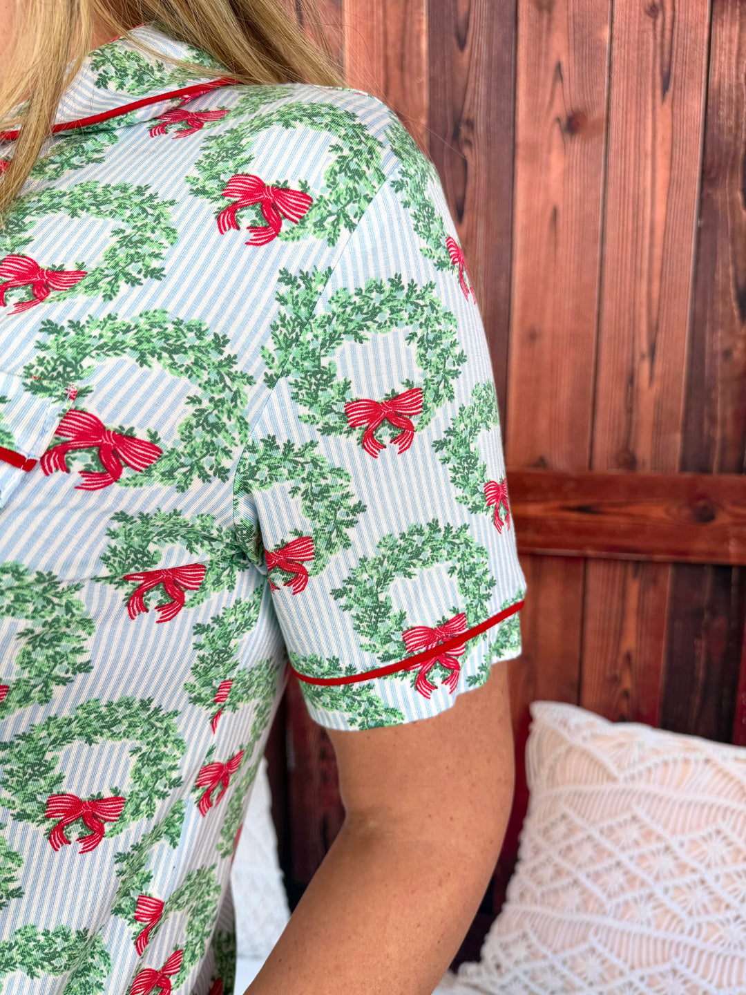 A close-up of a person wearing a short-sleeve pajama top with a green wreath and red bow holiday print on a white and light blue striped background, with red piping trim.