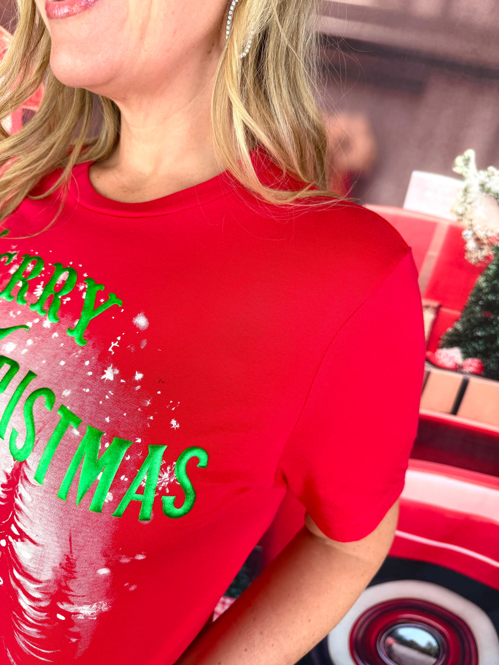 A close-up of a woman wearing a bright red short-sleeve t-shirt with 'MERRY CHRISTMAS' printed in green across the chest, set against a festive red and brown holiday background.
