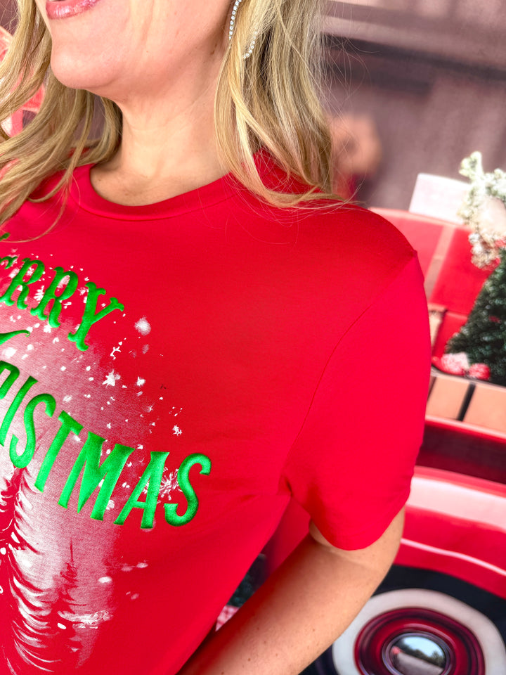 A close-up of a woman wearing a bright red short-sleeve t-shirt with 'MERRY CHRISTMAS' printed in green across the chest, set against a festive red and brown holiday background.