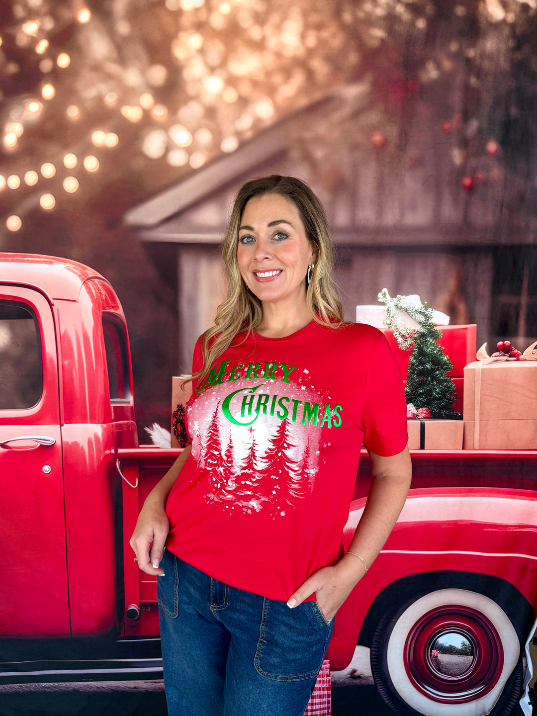 A woman wearing a red graphic t-shirt with 'Merry Christmas' and pine trees printed on the front, standing next to a vintage red pickup truck with holiday decorations in the background.