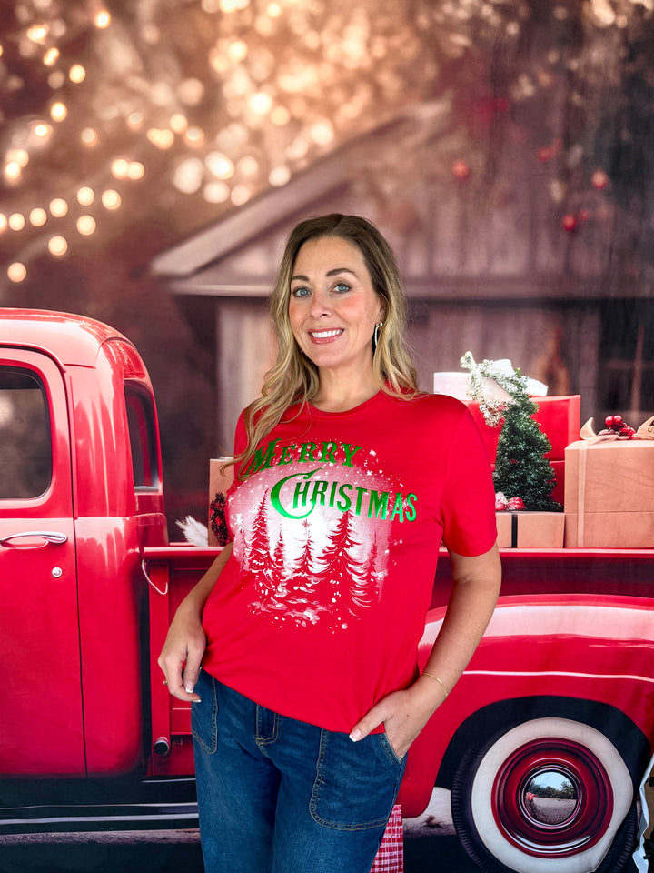 A woman wearing a red graphic t-shirt with 'Merry Christmas' and pine trees printed on the front, standing next to a vintage red pickup truck with holiday decorations in the background.