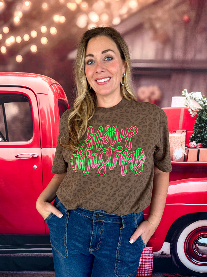 A woman wearing a brown leopard-print t-shirt with 'Merry Christmas' written in white and green script, paired with blue jeans, posed in front of a red vintage truck with holiday decorations.
