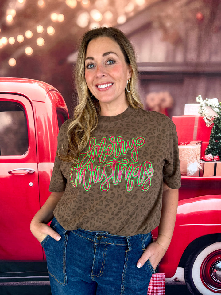 A woman wearing a brown leopard-print t-shirt with 'Merry Christmas' written in green and gold script, paired with blue jeans, standing in front of a red vintage truck with holiday decorations.