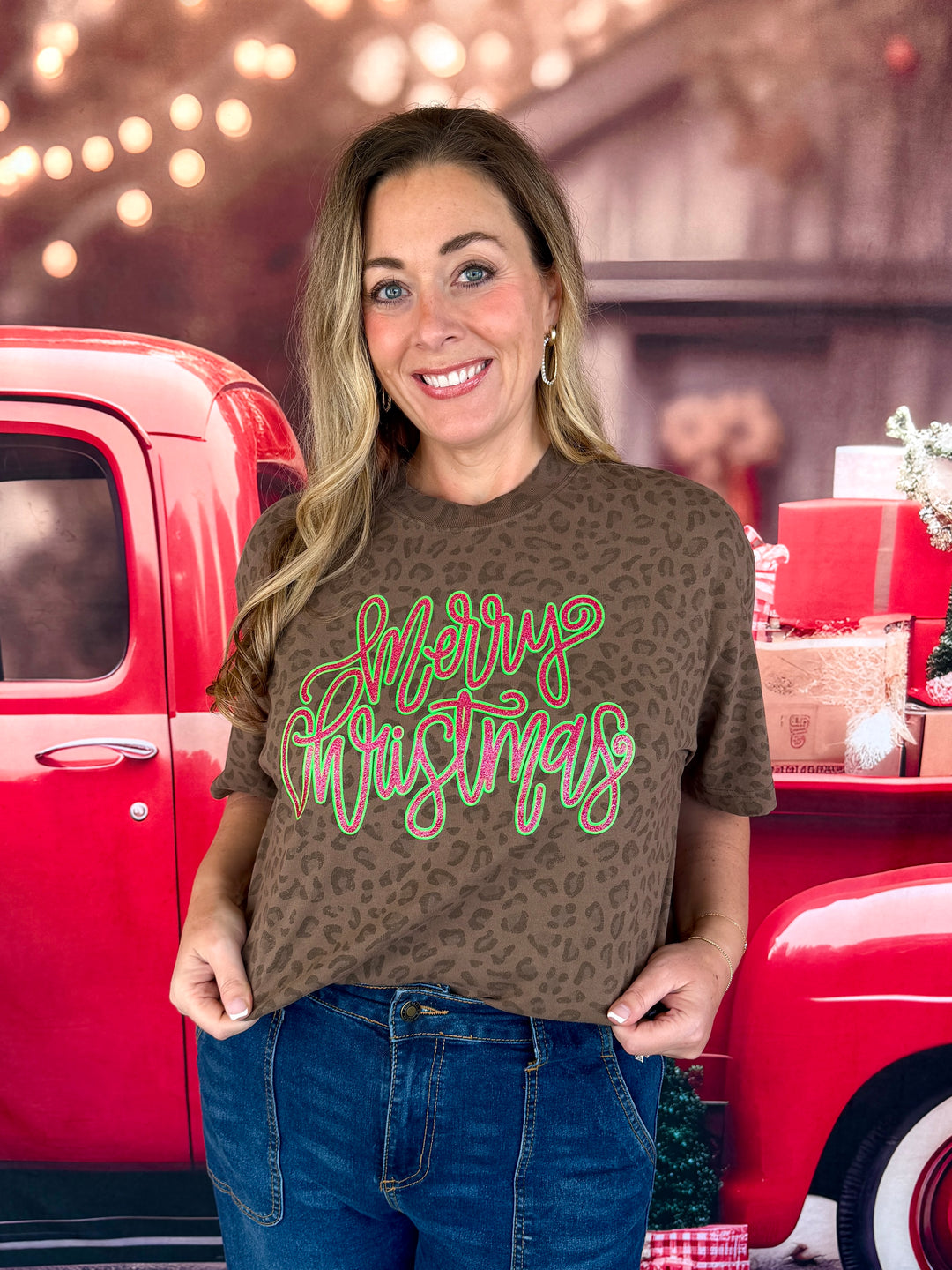 A woman wearing a brown leopard-print t-shirt with a 'Merry Christmas' graphic in green and pink script, standing in front of a red pickup truck decorated for the holidays.