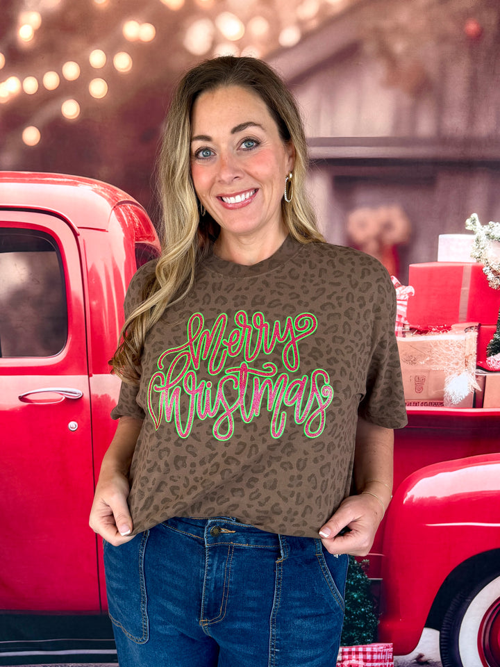 A woman wearing a brown leopard-print t-shirt with a 'Merry Christmas' graphic in green and pink script, standing in front of a red pickup truck decorated for the holidays.