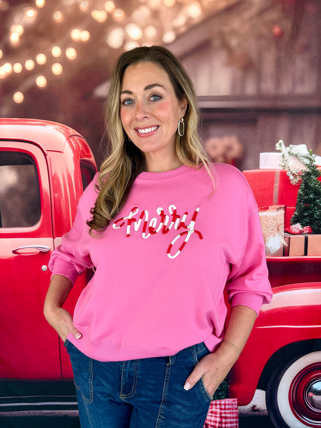 A woman wearing a bright pink crewneck sweatshirt with 'Merry' written in red and white cursive across the chest, standing in front of a red vintage pickup truck with holiday decorations.