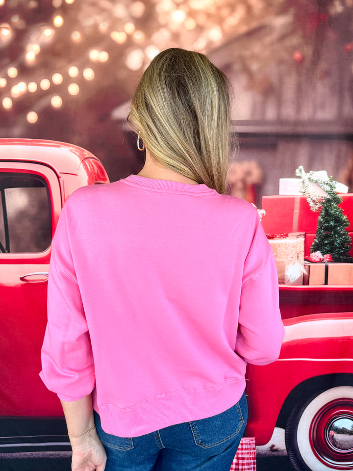 A woman viewed from behind wearing a bright pink crewneck sweatshirt and blue jeans, standing next to a red vintage pickup truck with holiday decorations in the background.