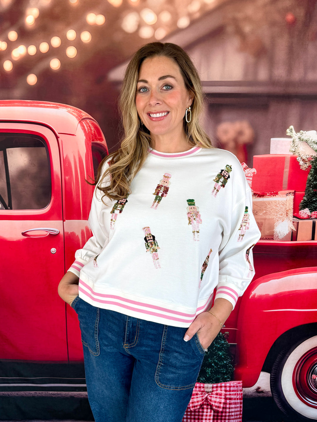 A woman wearing a white crewneck sweatshirt with pink stripes and nutcracker prints, paired with blue jeans, standing in front of a red vintage pickup truck decorated for the holidays.