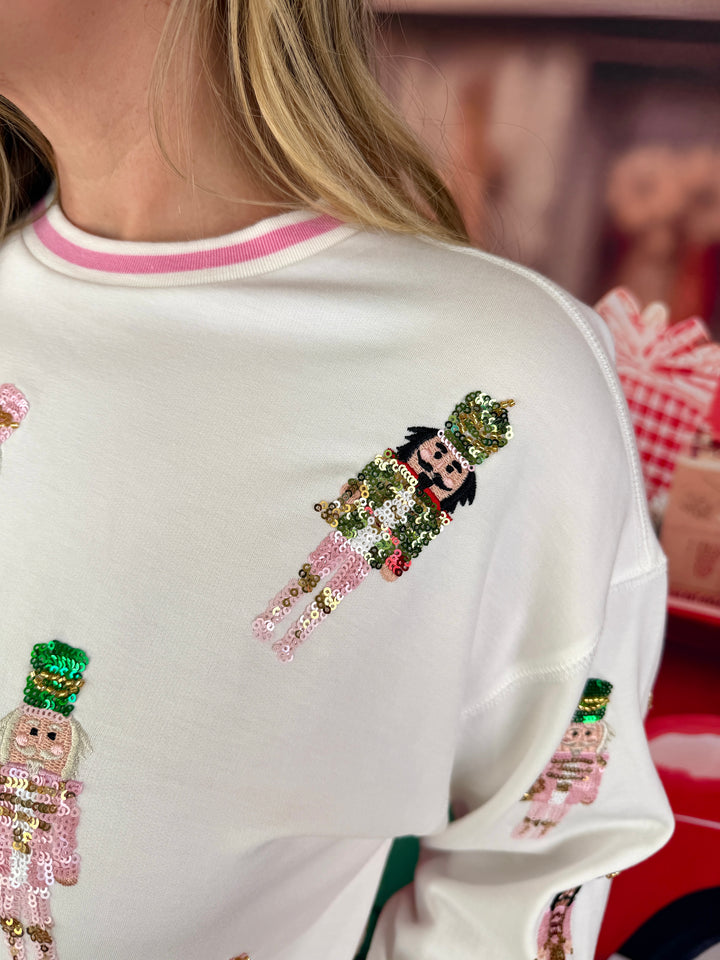 A close-up of a person wearing a white crewneck sweatshirt with pink trim and sequined nutcracker figures on the front.