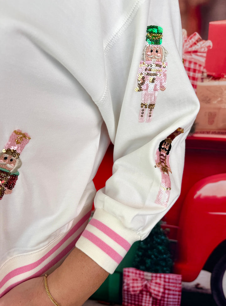 A close-up of a person wearing a white ribbed jacket with pink stripes on the cuffs and pockets, decorated with three sequined nutcracker figures on the sleeves and chest.