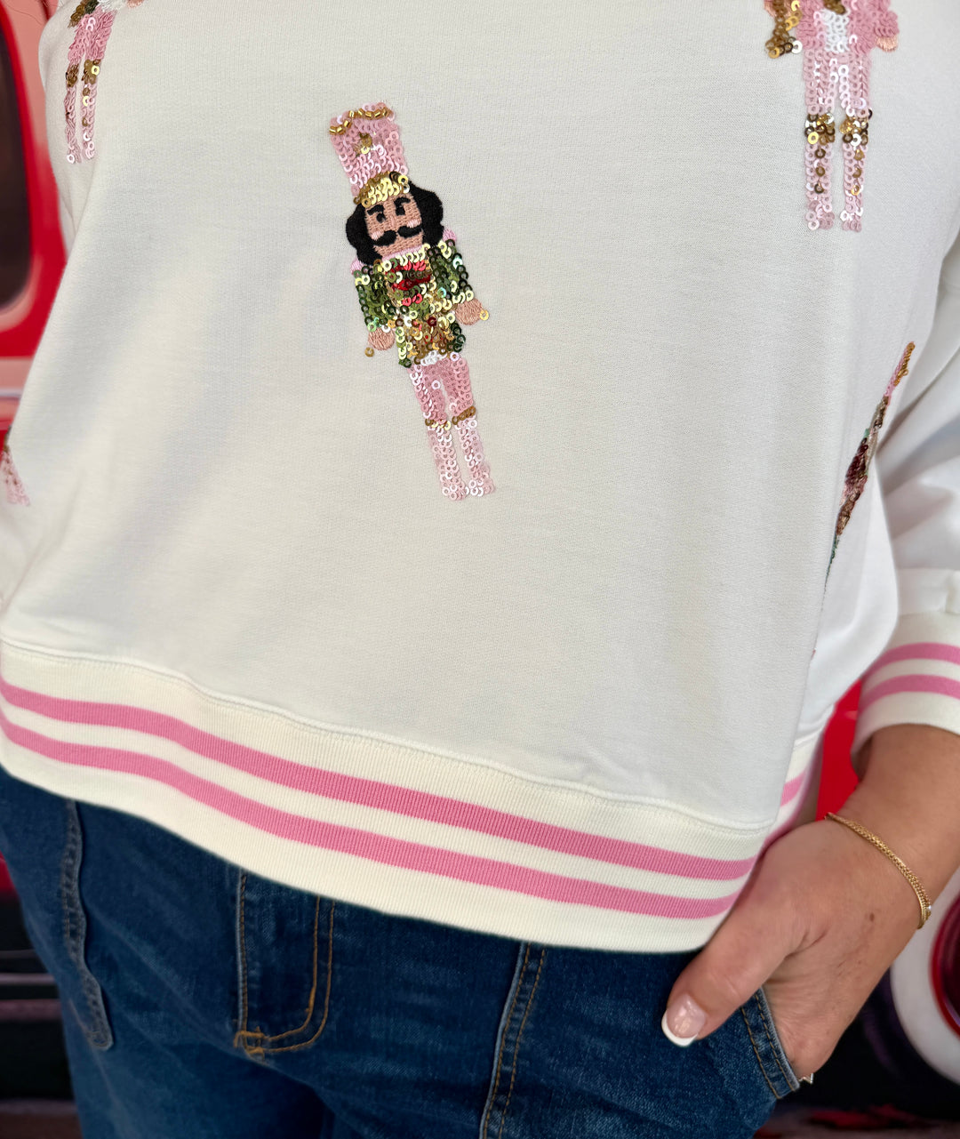 A close-up of a person wearing a white sweatshirt with pink and gold sequined nutcracker graphics and pink striped ribbed cuffs and hem, paired with blue jeans.