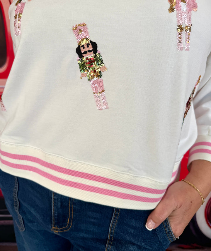 A close-up of a person wearing a white sweatshirt with pink and gold sequined nutcracker graphics and pink striped ribbed cuffs and hem, paired with blue jeans.