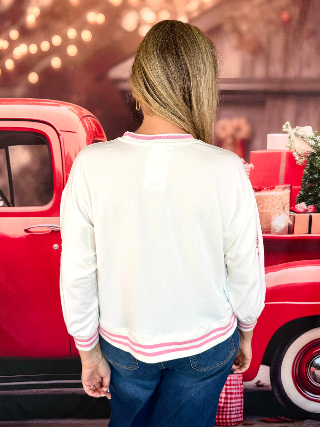 A woman viewed from behind wearing a white long-sleeve bomber-style top with pink stripes on the collar, cuffs, and hem, standing in front of a red vintage truck.