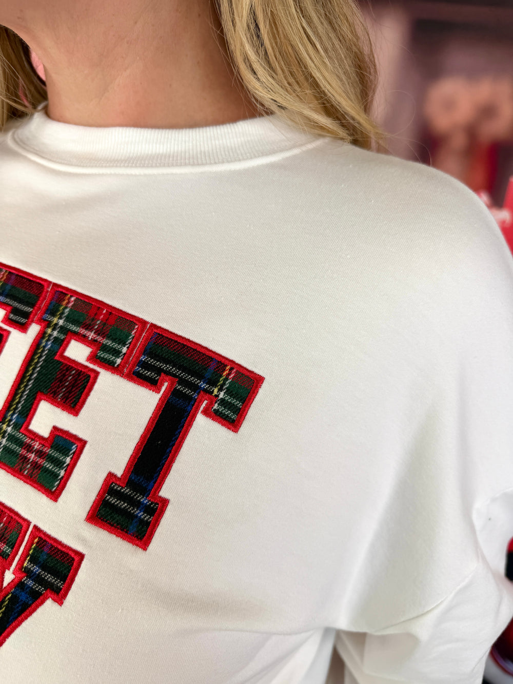 Close-up of a person wearing a white crewneck sweatshirt with large varsity-style letters outlined in red and filled with a green tartan plaid pattern.