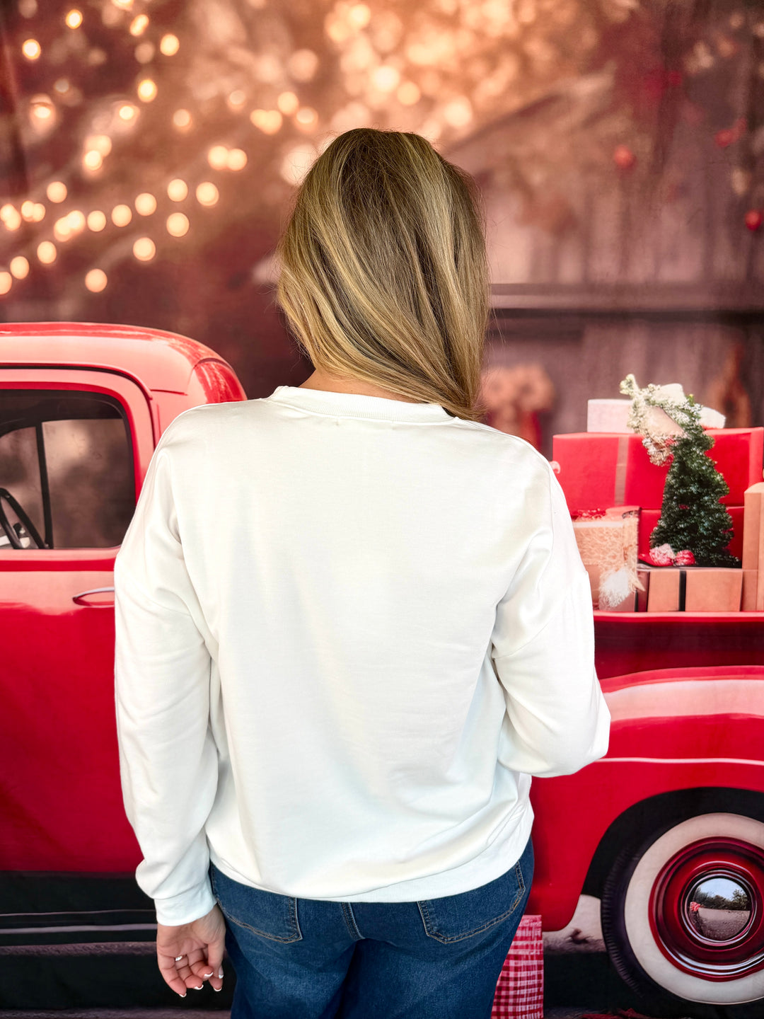 A woman viewed from behind wearing a white long-sleeve top and blue jeans, standing next to a red vintage truck decorated with holiday gifts and a small Christmas tree.