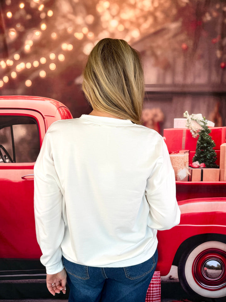 A woman viewed from behind wearing a white long-sleeve top and blue jeans, standing next to a red vintage truck decorated with holiday gifts and a small Christmas tree.