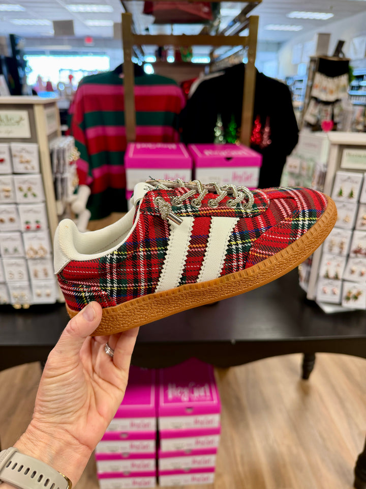 A hand holds a low-top sneaker with a red, green, and black tartan plaid pattern, featuring three white side stripes, white laces, and a brown gum sole, set against a retail store background.