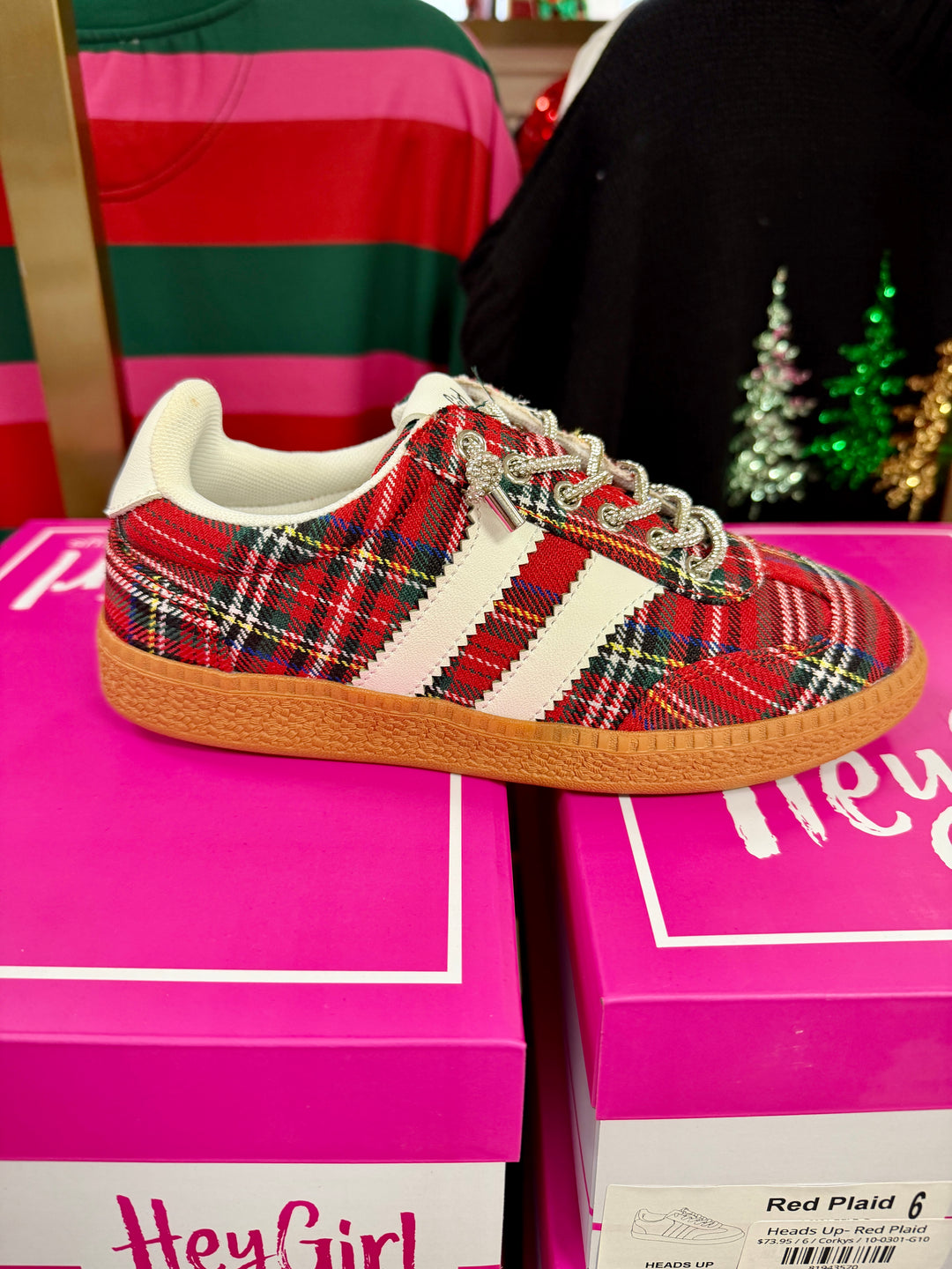 A red tartan plaid Gazelle sneaker with white side stripes and a gum sole, displayed on top of pink Hey Girl shoeboxes.