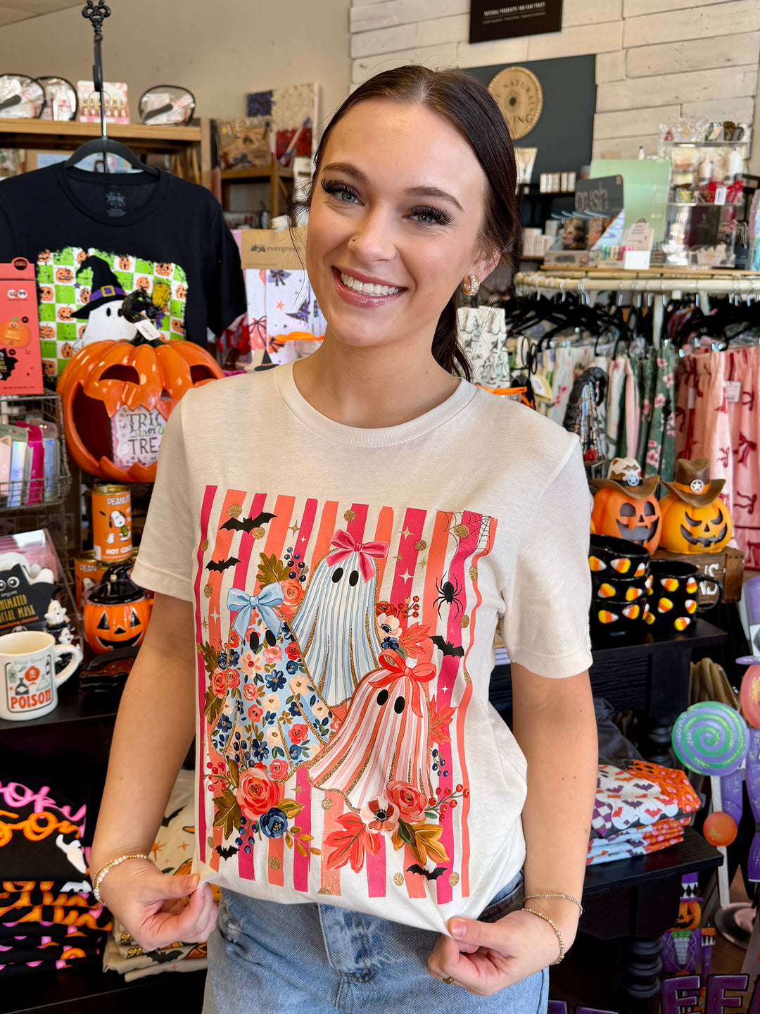 A woman wearing a cream-colored t-shirt with a colorful Halloween graphic print featuring ghosts, pumpkins, and stripes, standing inside a retail store.