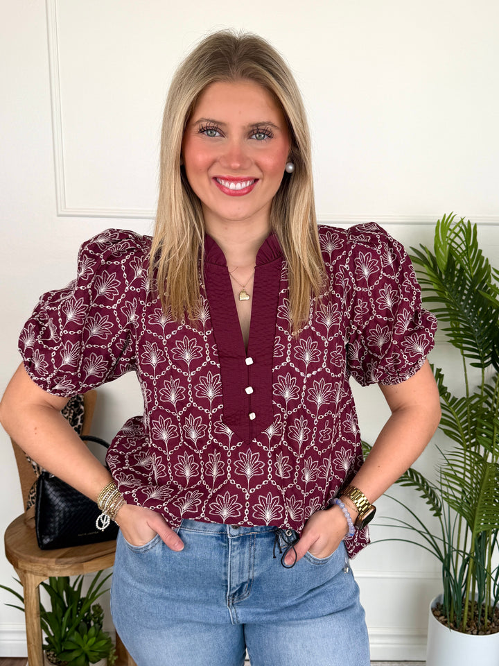 A smiling blonde woman wearing a burgundy and white patterned puff-sleeve blouse with a buttoned V-neck, paired with light blue jeans, standing indoors against a white wall with potted plants.