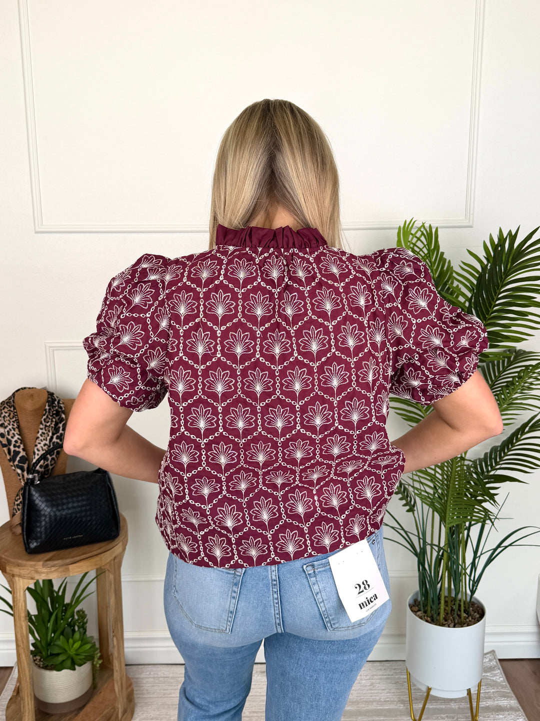 A woman viewed from behind wearing a burgundy short-sleeve blouse with a white geometric pattern and light blue jeans, standing in a room with indoor plants and a wooden side table.