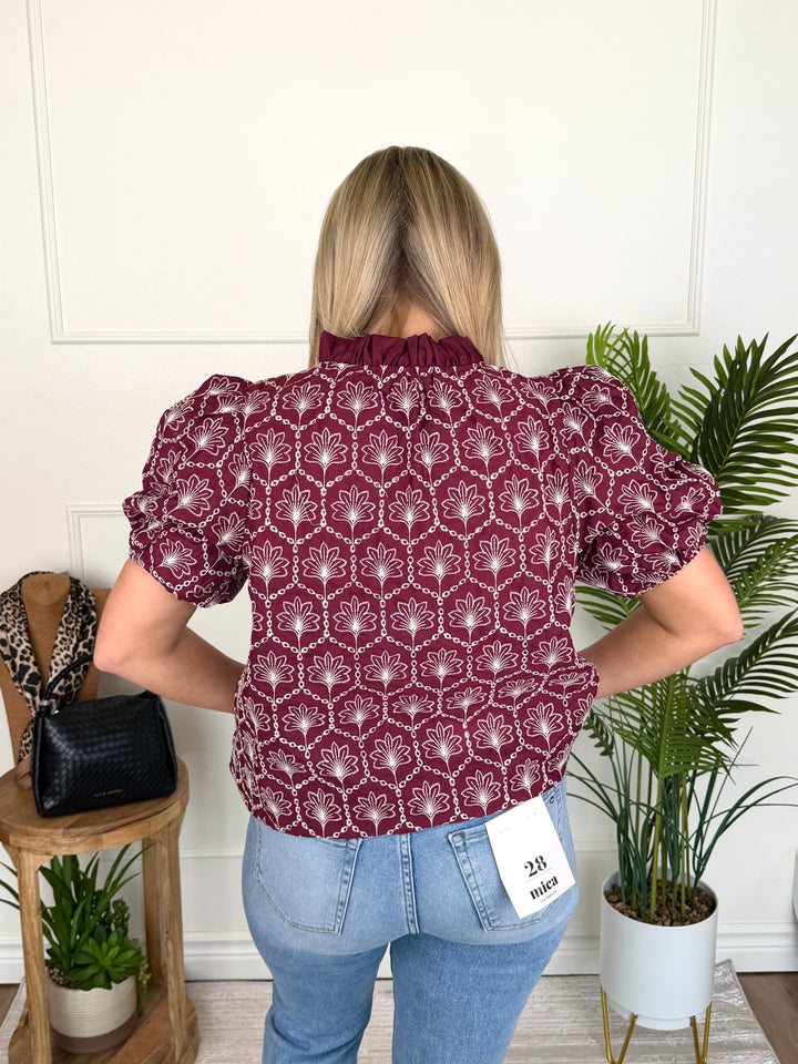 A woman viewed from behind wearing a burgundy short-sleeve blouse with a white geometric pattern and light blue jeans, standing in a room with indoor plants and a wooden side table.