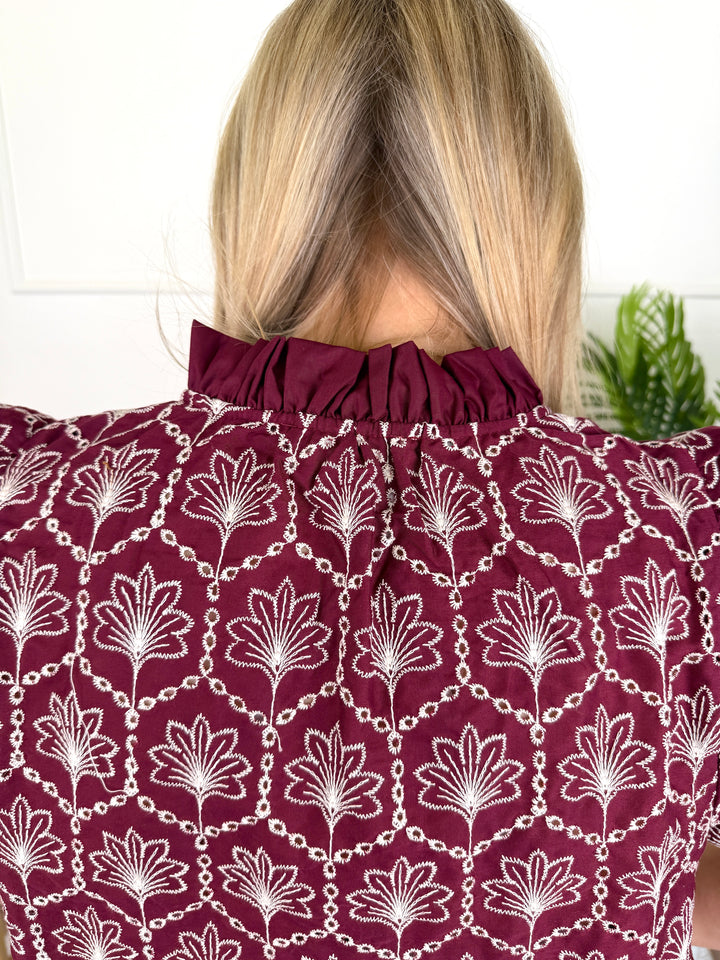 Back view of a woman wearing a maroon blouse with a white floral and chain pattern, featuring a gathered high neck.