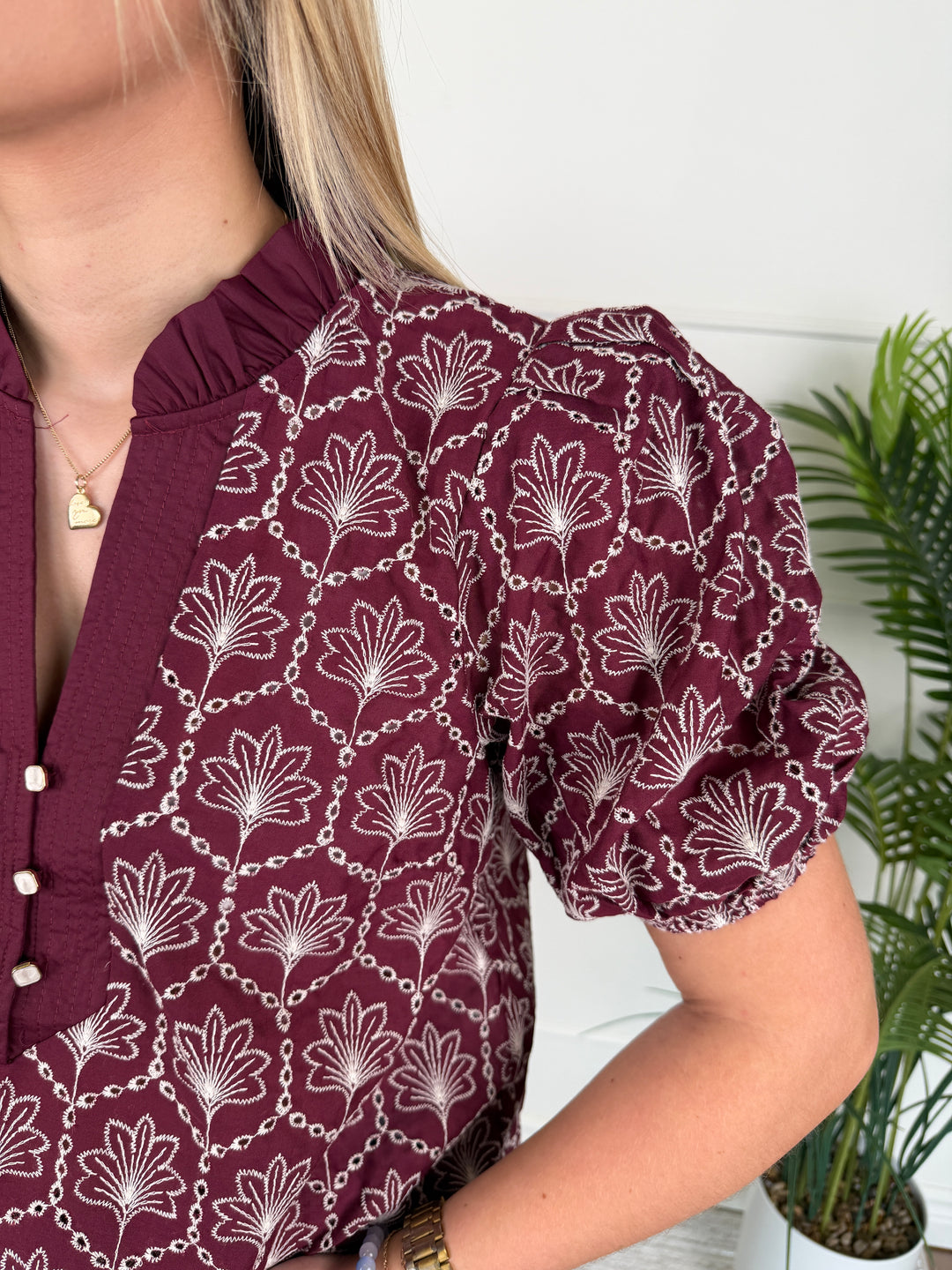 A close-up of a woman wearing a burgundy blouse with a white chain-link and floral pattern, featuring puff sleeves and a ruffled neckline.