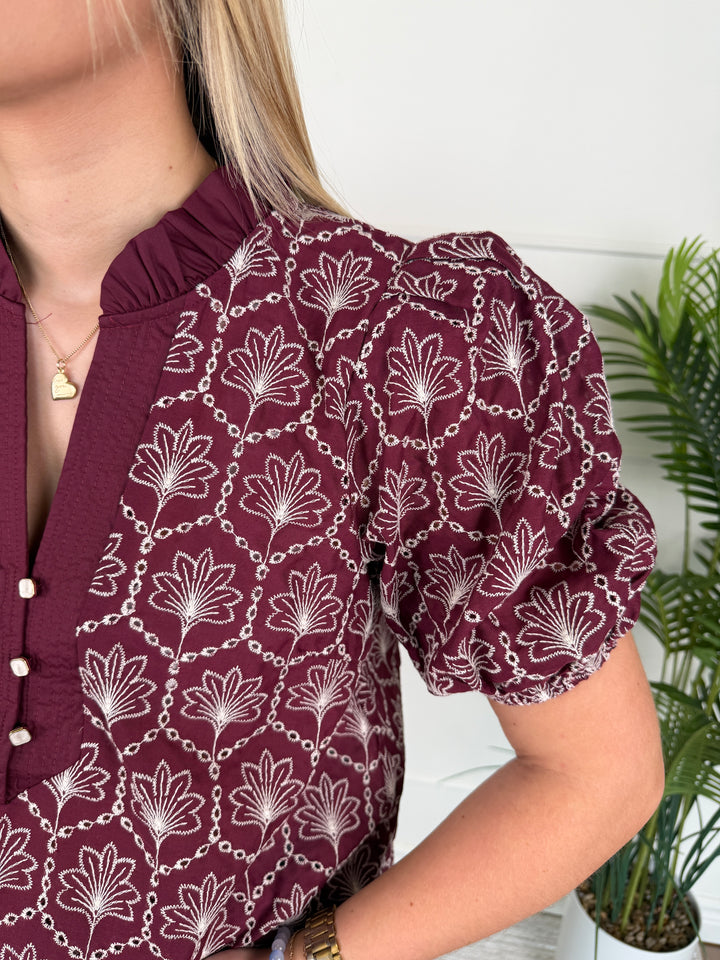 A close-up of a woman wearing a burgundy blouse with a white chain-link and floral pattern, featuring puff sleeves and a ruffled neckline.