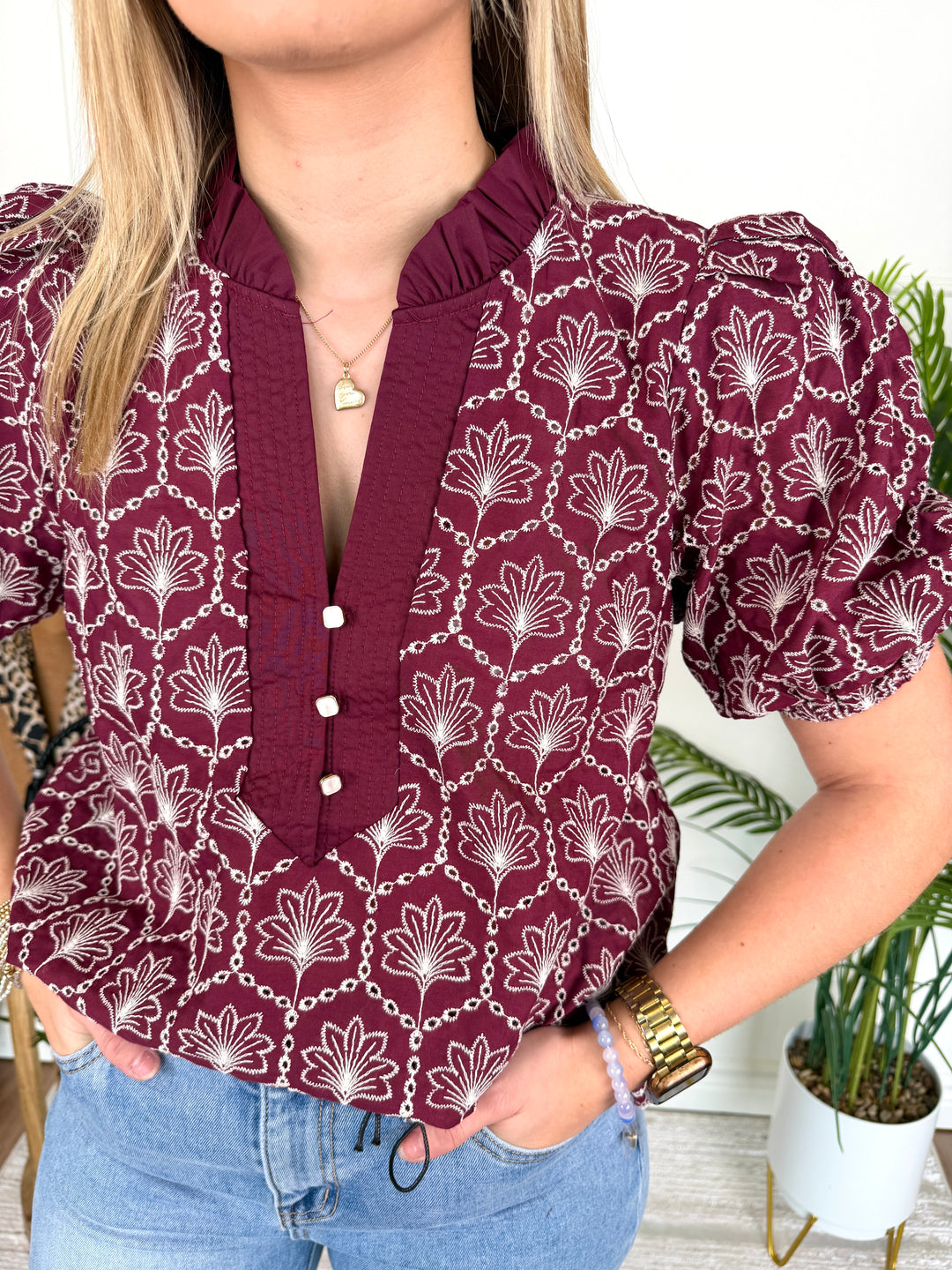 A woman wearing a burgundy short-sleeve blouse with a white floral and chain pattern, paired with light blue jeans. The blouse features a ruffled high collar and a buttoned front placket.