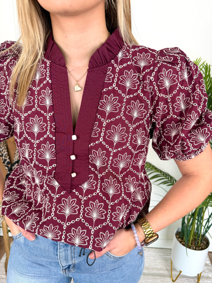 A woman wearing a burgundy short-sleeve blouse with a white floral and chain pattern, paired with light blue jeans. The blouse features a ruffled high collar and a buttoned front placket.