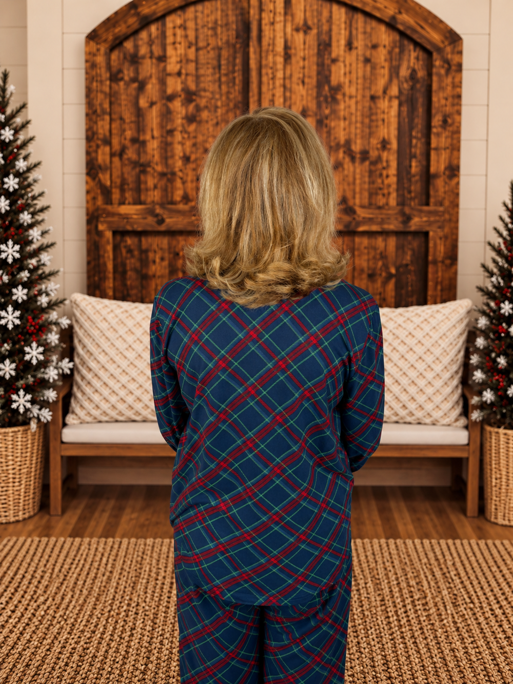 A woman viewed from behind wearing a navy blue pajama set with a red and green plaid pattern, standing in a cozy room with a wooden bench, Christmas trees, and a large rustic wooden door.