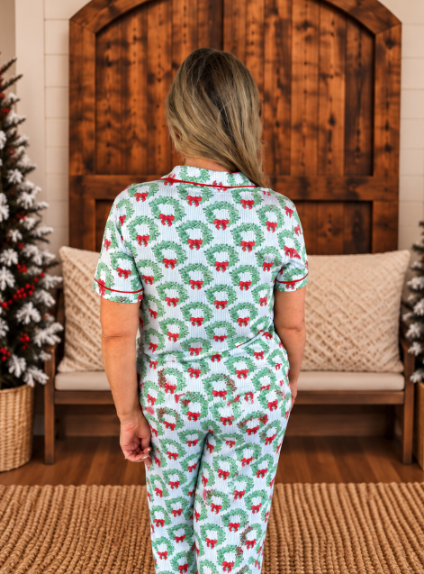 A woman viewed from behind wearing a matching short-sleeve pajama set with a green wreath and red bow holiday print, standing in a decorated room with a wooden bench and Christmas trees.