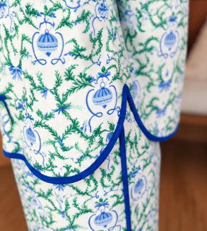 A close-up of a patterned fabric item, likely a blanket or towel, featuring a white background with a blue and green toile-style print and blue piping.